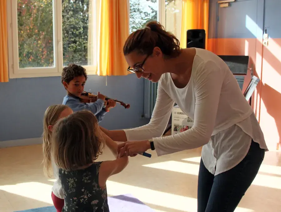 Image montrant une professeure et 3 jeunes enfants jouant du violon. La professeure aide une petite fille à placer son archet sur son violon.