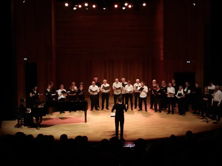 Photo montrant un choeur d'adultes chantant sur une scène dirigé  par une cheffe de choeurs et accompagné au piano