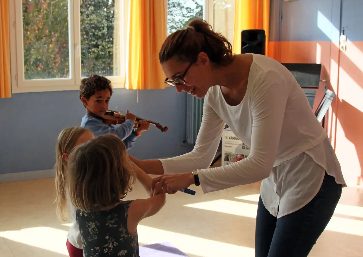 Image montrant une professeure et 3 jeunes enfants jouant du violon. La professeure aide une petite fille à placer son archet sur son violon.