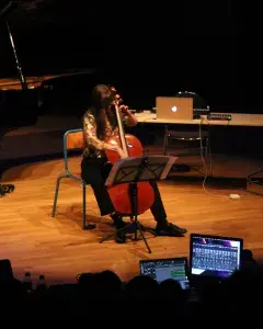 Photo montrant une musicienne jouant du violoncelle sur une scène devant l e public et un ordinateur passant de la musique électronique