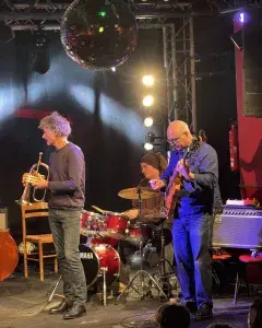 Photo montrant trois musiciens de jazz jouant sur une scène (trompette, batterie, guitare).
