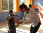 Image montrant une professeure et 3 jeunes enfants jouant du violon. La professeure aide une petite fille à placer son archet sur son violon.