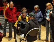 Photo montrant une femme montrant à un monsieur assis comment utiliser un accordéon. Ils sont entourés par 4 autres personnes qui écoutent la démonstration.