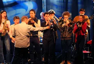 Photo montrant un groupe de 10 musiciens jazz jouant sur une scène en concert (saxophone, trombone, trompette) 