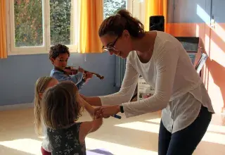 Image montrant une professeure et 3 jeunes enfants jouant du violon. La professeure aide une petite fille à placer son archet sur son violon.