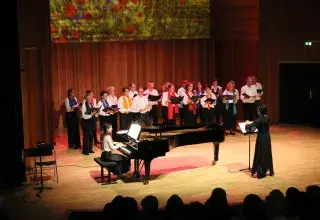 Photo montrant une vingtaine de choriste chantant sur scène accompagnée par une pianiste et dirigés par une cheffe de choeur.