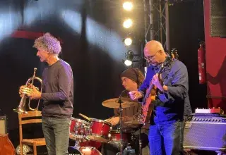 Photo montrant trois musiciens de jazz jouant sur une scène (trompette, batterie, guitare). 