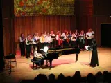Photo montrant une vingtaine de choriste chantant sur scène accompagnée par une pianiste et dirigés par une cheffe de choeur.