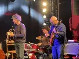 Photo montrant trois musiciens de jazz jouant sur une scène (trompette, batterie, guitare). 